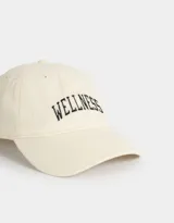 Gorra deportiva color crudo, de tela canvas con bordado frontal de la palabra "Wellness" en letras negras estilo universitario. Tiene visera curva y tira ajustable trasera con hebilla metálica.