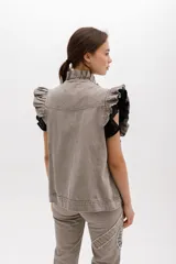 Chaleco de denim color topo o gris claro lavado, con volados estructurados en los hombros y a lo largo del frente. Es de corte recto, sin mangas, con cierre frontal de botones metálicos color cobre y bolsillos laterales.