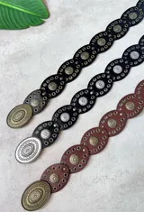 Cinto ancho de cuero con diseño de eslabones ovalados, cada uno adornado con múltiples ojales metálicos y un dije central con relieve floral. Se presenta en tres combinaciones de color: negro con herrajes plateados, negro con herrajes dorados y marrón con herrajes dorados. El cierre es una hebilla ovalada grande con relieve floral.