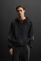 Cazadora con capucha confeccionada en tejido técnico ligero y elástico en todas las direcciones, color negro con detalles en gris. Cuenta con cuello con capucha ajustable, manga larga, bolsillos con cierre de cremallera en cadera, acabados con elástico y cierre frontal de cremallera.