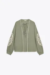 Blusa verde oliva de algodón con cuello redondo y escote en pico con cordones y borlas. Tiene manga larga con bordados florales en los hombros, jaretas y entredoses delanteros. Cierre frontal con botones.
