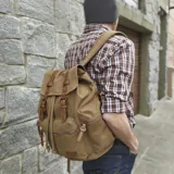Mochila de lona color marrón claro con detalles en cuero marrón. Tiene tapa con hebillas, cierre ajustable en la parte superior, bolsillos frontales y laterales con cierre, bolsillos internos, espacio para laptop, bolsillo trasero con cierre, correa de mano y correas regulables.