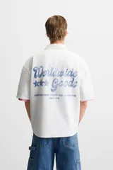 Camisa blanca de algodón con estructura canvas, corte boxy fit, cuello bowling y manga corta. Presenta bordado en azul claro con texto cursivo en el pecho y botones frontales.