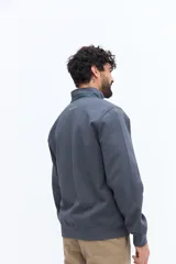 Campera de algodón gris azulado con cierre frontal completo y cuello alto. Presenta detalles de tela técnica azul verdoso en la parte superior del pecho y hombros, y un parche negro con texto blanco en el pecho izquierdo.