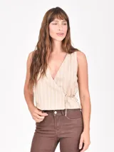 Chaleco sin mangas de color beige con rayas verticales blancas. Presenta un diseño cruzado tipo wrap con escote en V y ajuste mediante lazo lateral.