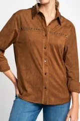 Camisa marrón de gamuza vegana con cuello clásico, cierre frontal con botones y mangas largas con puños abotonados. Presenta detalles de tachas metálicas en cuello y charreteras.