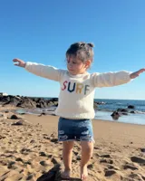 Sweater tejido a mano en lana merino color crudo con cuello redondo y mangas largas. Presenta la palabra "SURF" bordada en el frente con letras de colores.