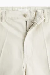 Jean tobillero de denim de algodón rígido color beige claro, con corte estándar de cadera a muslo y piernas con volumen de diseño curvo que se ciñen en el bajo. Presenta tiro alto, cierre con botón, bolsillos al bies, bolsillos decorativos traseros y pliegues delanteros y traseros.