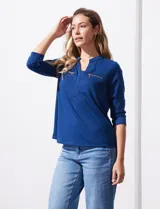Blusa blanca de poliéster y elastano, con cuello a la base, escote en V y mangas tres cuartos. Presenta dos bolsillos frontales con cierre metálico dorado.