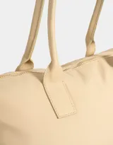 Bolso tipo shopper color beige con doble asa corta de hombro, cierre superior y tag de goma con logo.