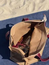 Bolso tipo tote de lona color beige con asas de cuero color bordó, cierre superior y bolsillo interno.