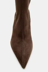 Bota alta de caña media en color marrón, confeccionada en material tipo gamuza. Presenta un diseño de punta fina, tacón alto y fino, y una tira decorativa con hebilla metálica en la parte superior de la caña. Incluye cierre lateral mediante cremallera.