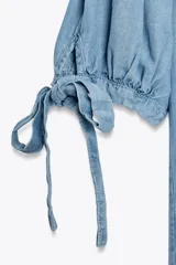 Camisa celeste de denim con cuello solapa, escote en pico, mangas abullonadas con lazos y cierre frontal con botones.