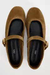 Zapato plano tipo bailarina color marrón de serraje, con escote redondo y cierre mediante hebilla en el empeine.