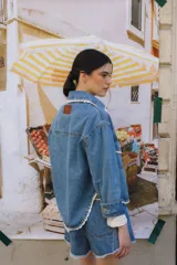 Campera de jean celeste con cuello camisero, cierre frontal con botones metálicos, bolsillos de parche en el pecho con solapa y detalle de puntilla blanca en cuello, solapas, bolsillos y ruedo.