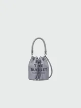 Bolso tipo bucket de cuero color gris con logo "The Bucket Marc Jacobs" estampado en negro. Tiene asa corta de cuerda trenzada y correa larga de cuero ajustable y removible.