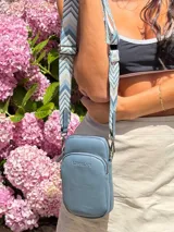 Bandolera pequeña tipo 'phone bag' de símil cuero color rosado pálido, con cierre de cremallera y correa ancha de tela con diseño de chevron en tonos claros.