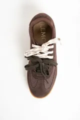 Zapatilla deportiva de perfil bajo, color marrón chocolate, con paneles de gamuza y malla texturizada. Presenta una entresuela de color crema y suela de goma color caramelo. Se ata con dos pares de cordones: uno blanco trenzado y otro verde oscuro.