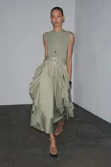 Vestido midi color verde oliva, de cuello redondo y sin mangas. Presenta un cinturón con hebilla metálica y detalle de volantes en la falda.