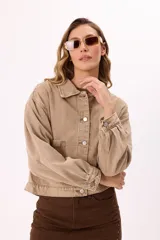 Campera de denim color beige, corte cropped, con cuello clásico, mangas largas abullonadas con puños abotonados, cierre frontal con botones metálicos y bolsillo de parche en el pecho.