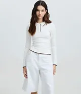 Blusa blanca de punto con cuello polo y mangas largas. Presenta detalles contrastantes en negro en el cuello, la abertura frontal y el dobladillo.