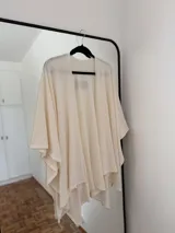 Ruana o poncho de tejido fino color crema o blanco roto, de corte amplio y asimétrico, sin cierres ni cuello definido.