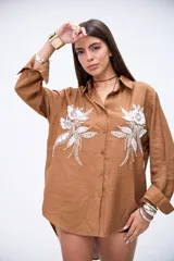 Camisa marrón de manga larga con puños anchos, cuello camisero y botones frontales. Presenta bordados florales en blanco con lentejuelas y mostacillas en la parte delantera.