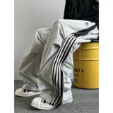 Pantalón deportivo color beige claro, estilo baggy o ancho, con cordón ajustable en la cintura y cierre completo impermeable (full zip) en los laterales. Presenta una franja vertical negra con tres líneas blancas a lo largo de la costura lateral, y un pequeño logo bordado en la parte superior del muslo.