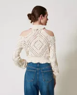 Sweater color crema de punto calado con motivo de rombos, cuello alto con volados y hombros descubiertos. Tiene mangas largas holgadas, ribetes acanalados, bajos festoneados y corte holgado.