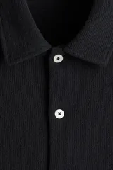 Camisa negra de punto con textura, de corte recto, manga corta, cuello inglés, tapeta lisa, canesú en la espalda y bajo recto con aberturas laterales.