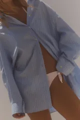 Camisa celeste con rayas verticales blancas, de corte amplio, cuello clásico, manga larga con puños abotonados, bolsillo de plastrón en el pecho y cierre frontal con botones.