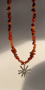 Collar de piedras de color naranja y dije de sol plateado. Las piedras son de diferentes tamaños y formas. El dije de sol es de bronce y tiene un diámetro de 2.5 cm.