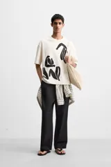 Camiseta color crudo de corte relaxed fit, cuello redondo y manga corta. Presenta un estampado abstracto con figuras humanas en color negro.
