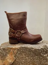 Bota corta de cuero color marrón, con taco cuadrado y punta redonda. Presenta una correa con hebillas y una argolla metálica decorativa alrededor del tobillo.