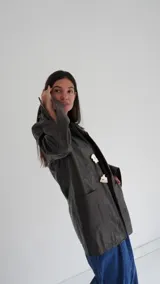 Blazer de cuero color gris oscuro, con aberturas laterales, aplique tipo pin desplegable bañado en bronce y botones de nácar.