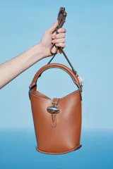 Cartera tipo balde color marrón con asa de mano rígida y correa bandolera ajustable y extraíble. Cierre con trabilla y adorno metálico dorado.
