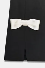 Vestido corto negro con cuello redondo y sin mangas. Presenta dos lazos blancos en la parte inferior delantera y aberturas frontales en el ruedo. Cierre trasero con cremallera metálica.