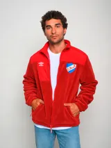 Campera polar roja con cierre frontal, cuello alto y bolsillos laterales. Presenta un panel liso en la parte superior del pecho y un bolsillo con cierre en el lado izquierdo. Incluye el logo de Umbro bordado en el pecho izquierdo y el escudo del Club Nacional de Football bordado en el pecho derecho.