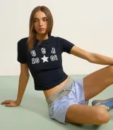 Remera baby look azul marino con estampado frontal de letras "USA" apiladas verticalmente sobre el número "20" y el número "08" separados por una estrella blanca.