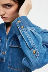 Camisa de denim azul de corte holgado, con cuello mao, tapeta de botones, hombros caídos, mangas largas con puños abotonados y dos bolsillos superiores con solapa.