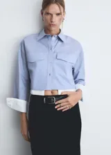 Camisa celeste de popelina de algodón con diseño crop, cuello camisero, cierre frontal con botones, dos bolsillos de solapa en la parte delantera, manga larga con puños abotonados y terminaciones en contraste blanco.