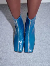 Botas de cuero charol negro con caña media y punta cuadrada. Cuentan con cierre trasero, tirador y bordado contrastante en el empeine. El taco es de madera y la suela de goma.