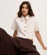 Camisa cropped de sarga color crudo, con cuello clásico, cierre frontal con botones dorados, manga corta, bolsillo de parche en el pecho y costuras a contraste. Confeccionada con algodón certificado.