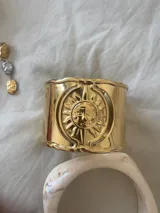 Brazalete ancho dorado con diseño de sol grabado en el centro.