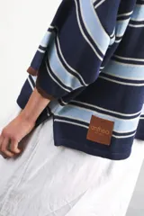 Remera de algodón y poliéster a rayas horizontales rojas y azul marino sobre fondo crudo. Tiene cuello tipo polo con botones, mangas amplias y corte oversized.