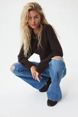 Jeans de denim azul medio con ligero efecto desgastado y roturas a la altura de las rodillas. Presenta un corte ajustado en muslos y acampanado desde la rodilla. El tiro es bajo, con pretina prolongada que se cierra con dos botones visibles. Incluye bolsillos delanteros tipo parche y bolsillos traseros.