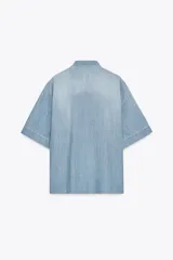 Camisa de denim celeste con cuello solapa y manga corta. Presenta detalle de volantes en el frente y bajo asimétrico con aberturas laterales.