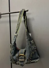 Cartera bandolera de denim azul con detalles de tachas y hebilla metálica.