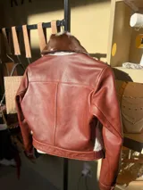 Campera de cuero color marrón rojizo (whisky) con detalles de piel de vaca (marrón y blanco) en el cuello y solapas. Se observa que está abierta, revelando un sweater de punto color crema con botones dorados debajo.