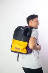 Mochila de nylon con diseño tricolor: negro en la parte superior, amarillo en el bolsillo frontal y morado en los laterales. Presenta el logo de la NBA en la parte superior y el logo de Los Angeles Lakers en el bolsillo frontal. La espalda es de tela mesh.